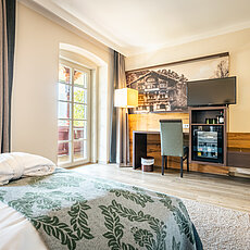 Gemütliches Hotelzimmer mit Bett, Schreibtisch, Fernseher, Minibar und Zugang zum Balkon bei Tageslicht
