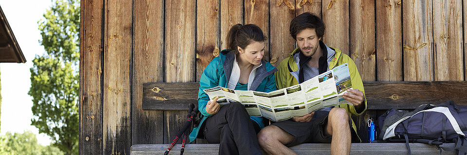 Wanderpaar liest eine Wanderkarte auf einer Bank vor einer Holzhütte