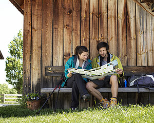 Wanderpaar liest eine Wanderkarte auf einer Bank vor einer Holzhütte
