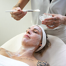 Frau erhält Gesichtsbehandlung mit weißer Creme, aufgetragen von Kosmetikerin mit Pinsel