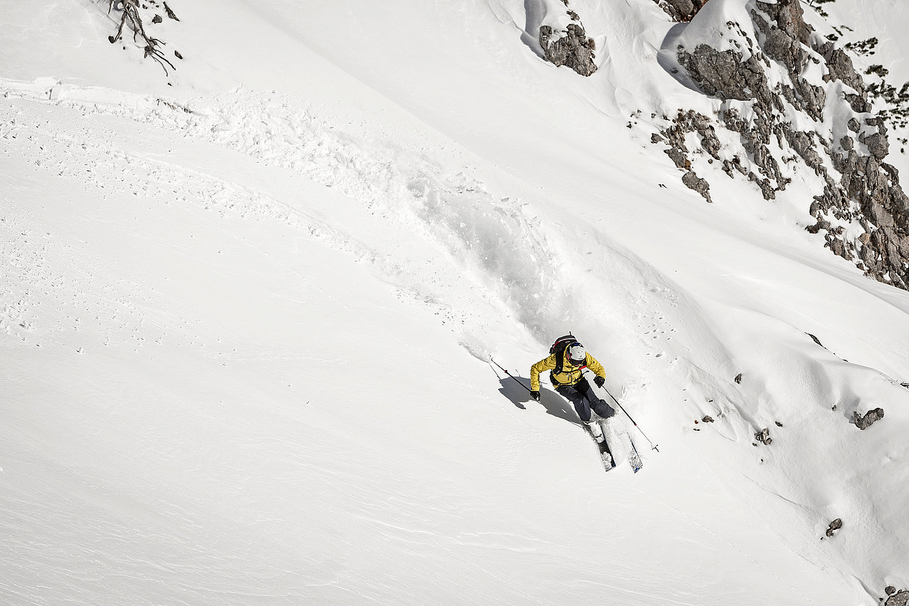 Backcountry skier in yellow jacket carving through fresh powder on a steep snowy mountain slope