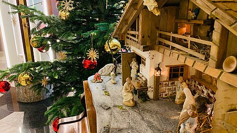 Weihnachtliche Krippenszene mit Figuren neben geschmücktem Baum mit roten und goldenen Kugeln