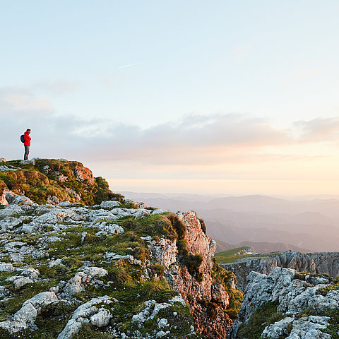 Wanderer in roter Jacke auf felsigem Berggipfel bei Sonnenaufgang mit Panoramablick ins Tal