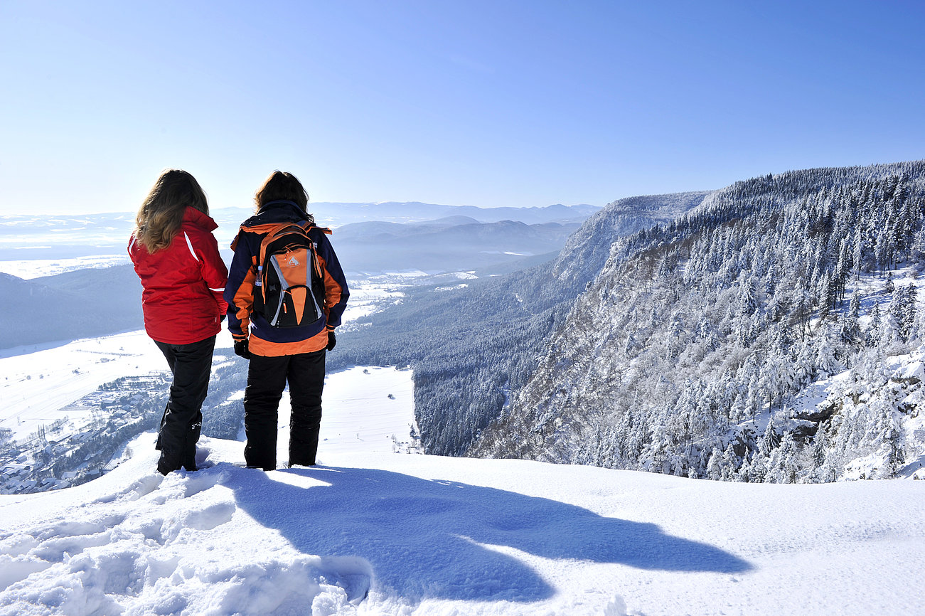 Zwei Wanderer blicken auf ein verschneites Bergtal unter klarem blauen Himmel