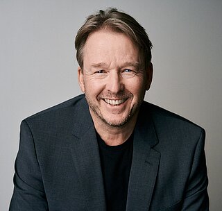 Lächelnder Mann in dunklem Blazer und schwarzem Shirt vor neutralem Hintergrund