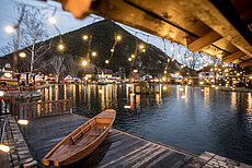 Gemütliches Dorf am See mit Holzboot, Lichterketten und bewaldetem Berg in der Abenddämmerung