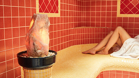 Leuchtende Himalaya-Salzlampe in gefliester Dampfsauna mit entspannter Person auf Bank