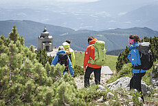 Group of hikers with backpacks walking down a mountain trail toward an alpine chapel