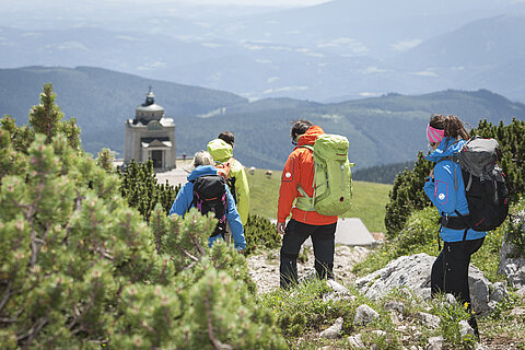 Group of hikers with backpacks walking down a mountain trail toward an alpine chapel