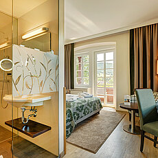 Cozy hotel room with open glass bathroom, balcony view, and work desk