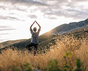 Frau macht Yoga-Baumhaltung im Grasfeld bei Sonnenaufgang mit Bergblick