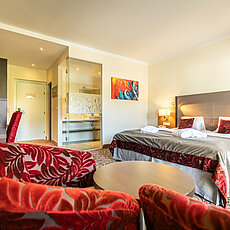 Modern hotel room with double bed, red armchairs, desk, and open bathroom area