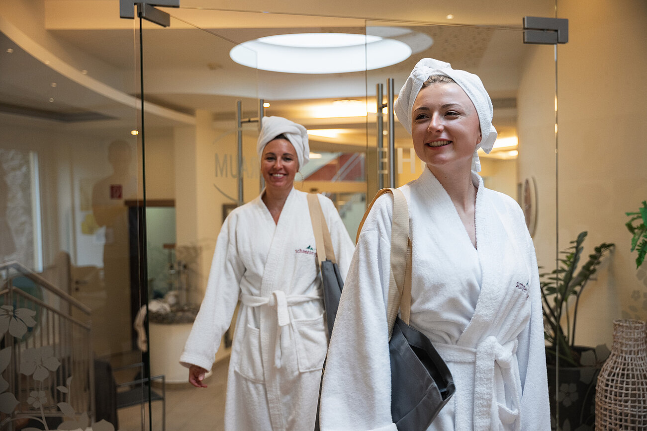 Two smiling women in white bathrobes and towels entering a wellness spa