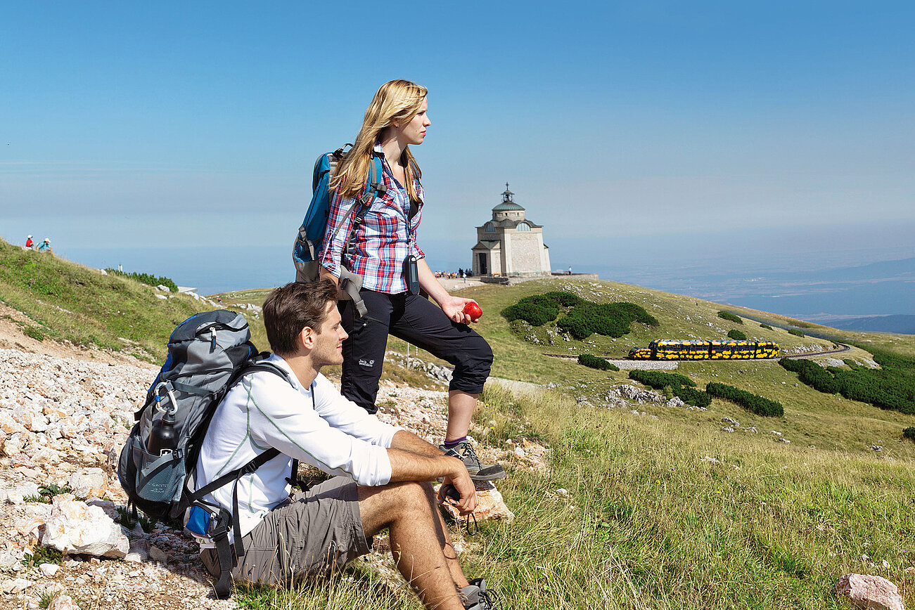 Wanderer rasten auf felsigem Hügel nahe einer Kapelle und einer Bergbahn im Sommer