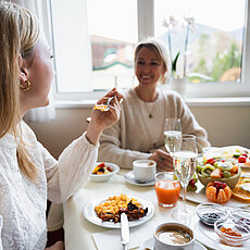 Zwei Frauen genießen ein Frühstück mit Kaffee, Saft, Obst und Gebäck an einem sonnigen Tisch