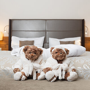 Zwei Teddybären in weißen Bademänteln auf einem Hotelbett mit Handtüchern und Kissen