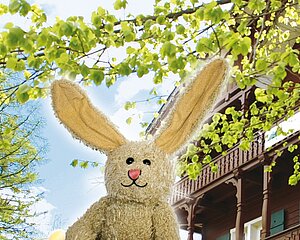 Plüsch-Osterhase mit langen Ohren vor belaubtem Baum und traditionellem Haus