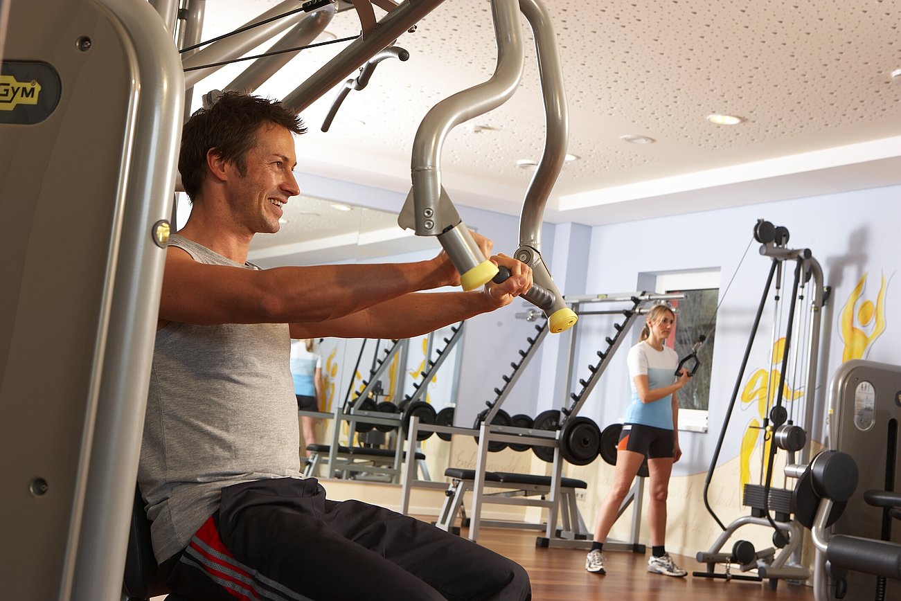 Mann trainiert an der Brustpresse in modernem Fitnessstudio, Frau im Hintergrund beim Training