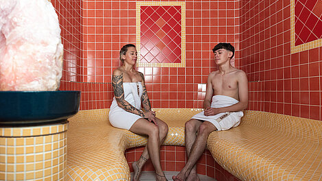 Zwei Personen entspannen auf gefliesten Bänken in einem roten und gelben Dampfbad