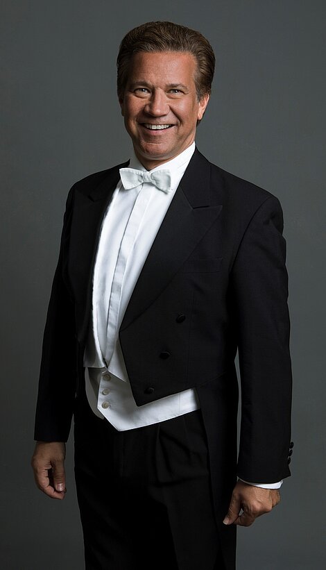 Smiling man in formal black tuxedo with white bow tie standing against gray background