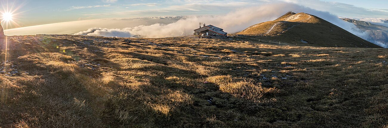 Berghütte auf alpiner Wiese im Sonnenaufgang mit Panoramablick und Wolken