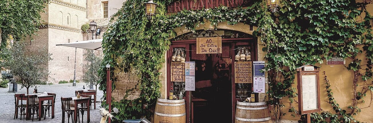 Charmante italienische Trattoria mit efeubewachsener Fassade, Außentischen und Vintage-Roller auf Kopfsteinpflaster