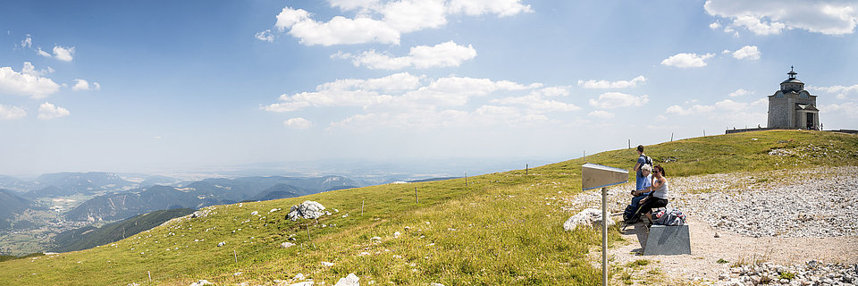 Wanderer rasten neben Kirche auf grasigem Hochplateau mit Panoramablick ins Tal