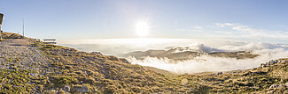 Berghütte auf felsigem Plateau mit Panoramablick auf Sonnenaufgang über Alpenwolken
