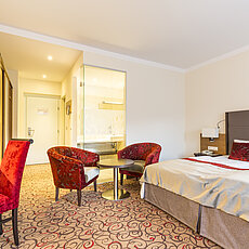 Modern hotel room with double bed, red armchairs, desk, and open bathroom area