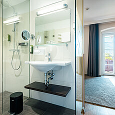 Modernes Hotelbad mit Glasdusche und Waschbecken neben hellem Zimmer mit Balkonblick