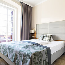 Helles modernes Hotelzimmer mit Doppelbett, grauem Kopfteil und großem Fenster mit Vorhängen