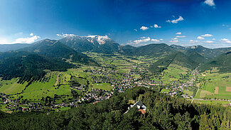 Panoramablick auf grünes Tal mit Stadt, umgeben von bewaldeten Bergen und schneebedeckten Gipfeln