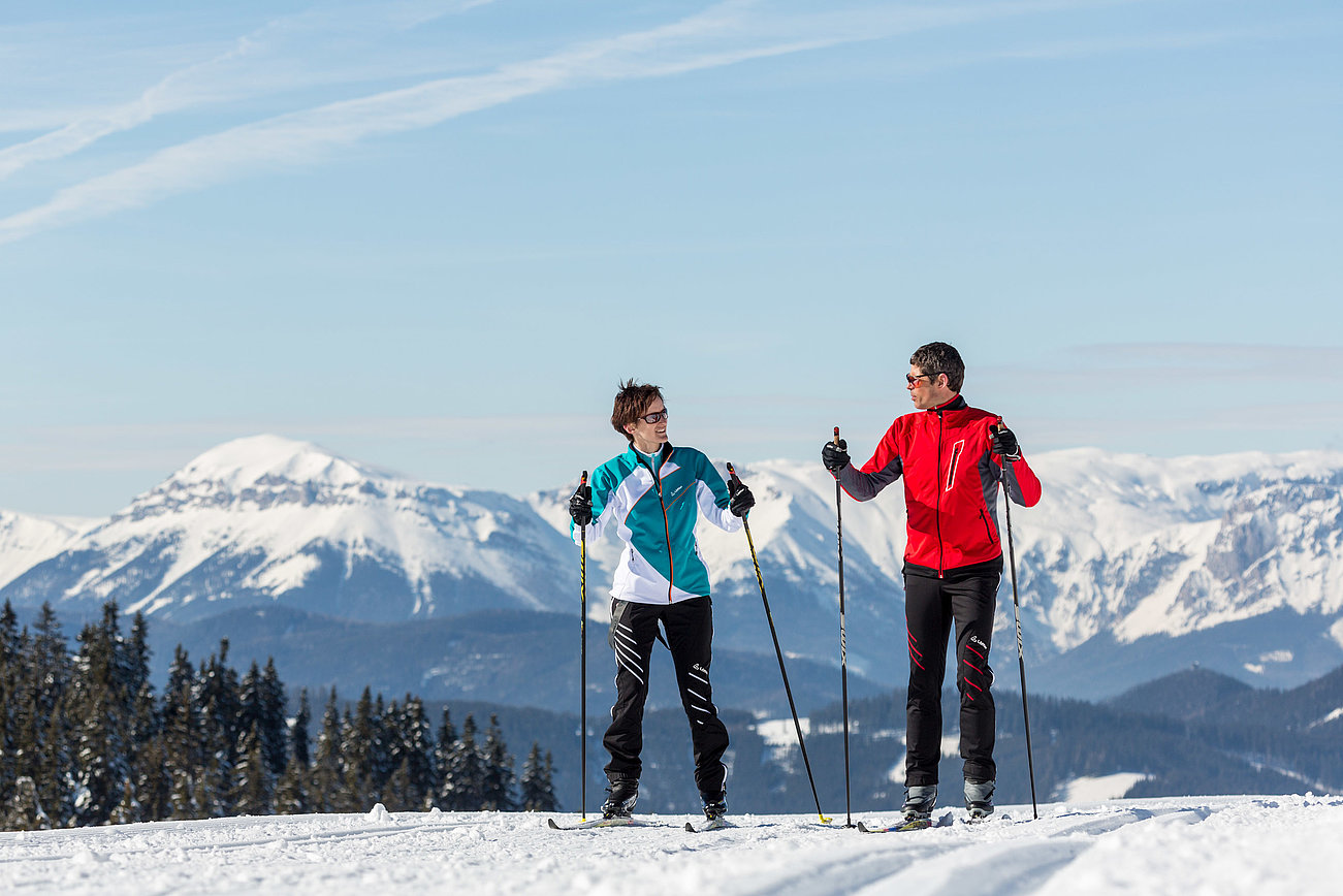 Zwei Langläufer unterhalten sich auf verschneiter Loipe vor Bergkulisse