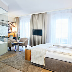 Modern hotel room with double bed, glass-walled bathtub, armchairs, and large windows with curtains