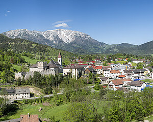 Malerisches Alpendorf mit Kirche und Burg, umgeben von grünen Hügeln und schneebedeckten Bergen