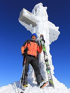 Bergsteiger mit Skiern neben schneebedecktem Gipfelkreuz unter klarem blauen Himmel