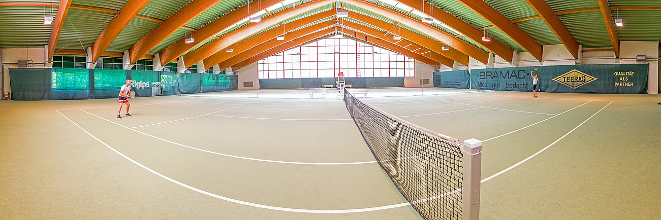 Two people playing tennis on an indoor court with arched wooden ceiling