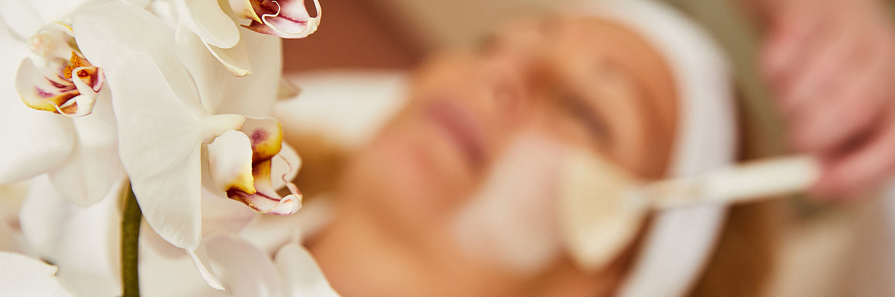 Weiße Orchideenblüten im Vordergrund mit Frau bei Gesichtsbehandlung im Hintergrund