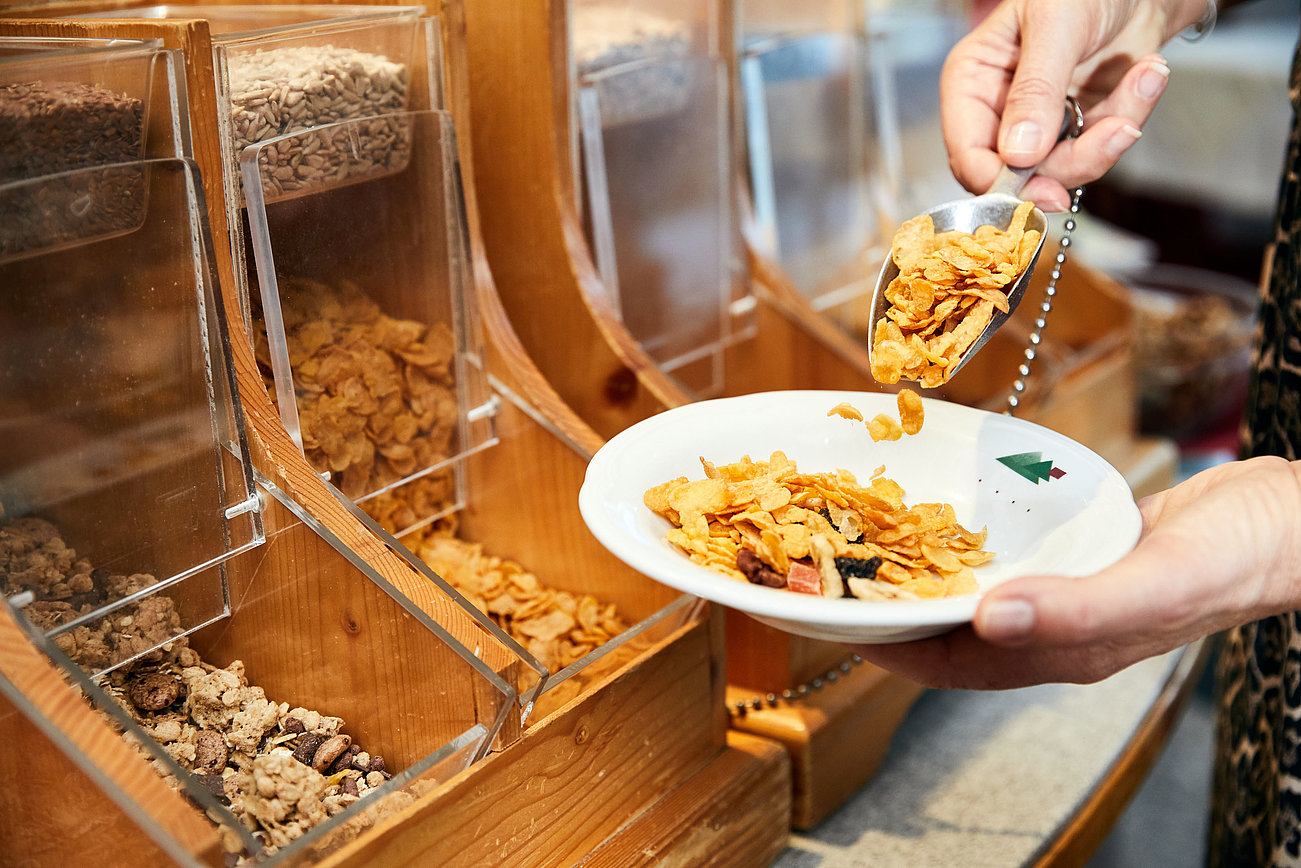Hand schöpft Cornflakes in eine Schüssel an einer Selbstbedienungs-Müslistation