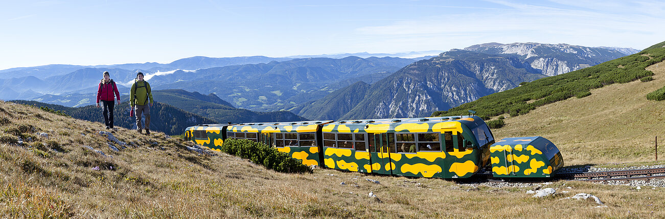 Zwei Wanderer gehen neben grün-gelbem Bergzug in den österreichischen Alpen