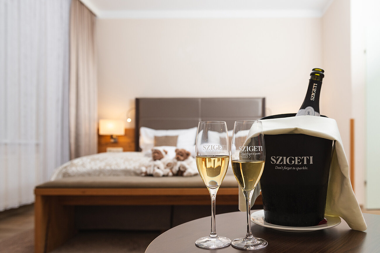 Zwei Gläser Sekt mit Szigeti-Flasche in einem Hotelzimmer mit Doppelbett