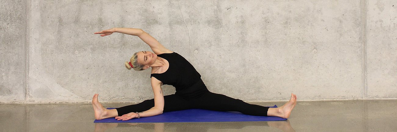 Frau macht sitzende Seitdehnung-Yoga-Pose auf blauer Matte vor Betonwand