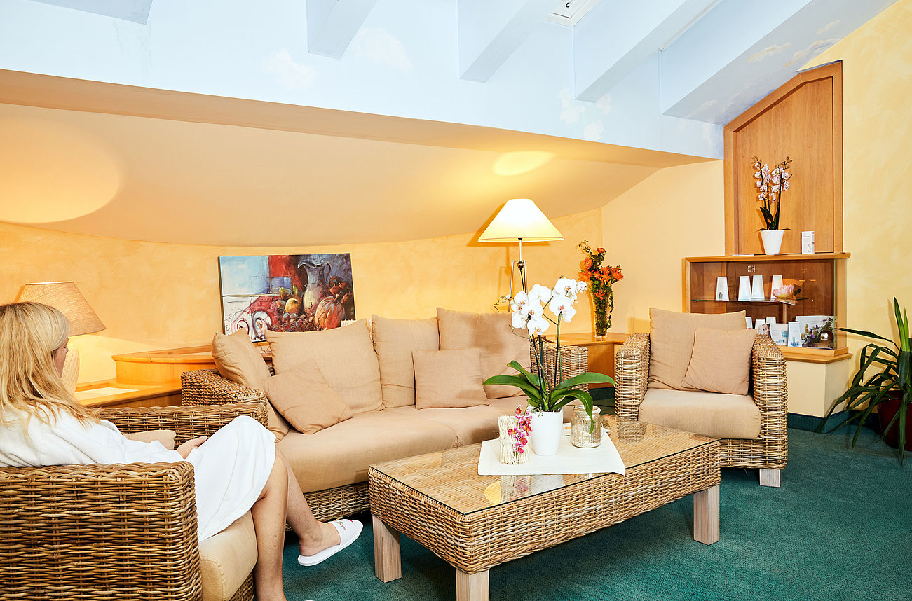 Gemütlicher Spa-Loungebereich mit Korbmöbeln, Orchideen auf Glastisch und Frau in Bademantel