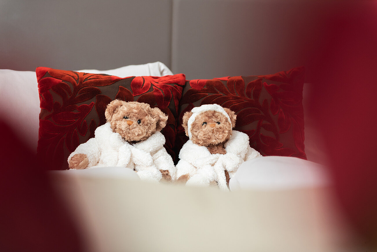Zwei Teddybären in weißen Bademänteln auf einem Bett mit roten Zierkissen