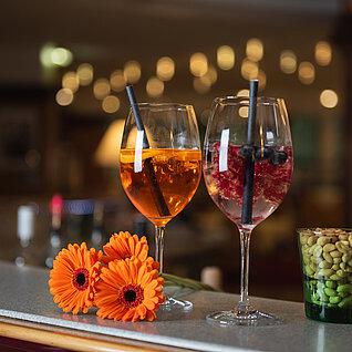 Zwei Cocktails mit Beeren und Eis auf einem Tresen mit Erdnüssen und orangefarbenen Gerbera-Blumen