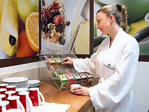 Frau im Bademantel wählt Tee am Hotel-Frühstücksbuffet mit roten Tassen und Fruchtdeko