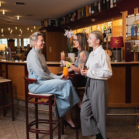 Drei Frauen unterhalten sich mit Cocktails an einer stilvollen Bar mit warmer Beleuchtung