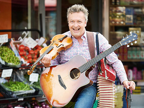 Lächelnder Straßenmusiker mit Gitarre, Akkordeon und Geige vor Gemüsemarkt