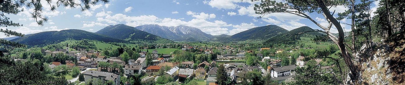 Panoramablick auf ein Alpendorf mit bewaldeten Hügeln und schneebedeckten Bergen im Frühling